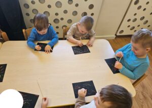 Dzieci siedzące przy stoliku, wydrapują patyczkami w czarnych kartkach.