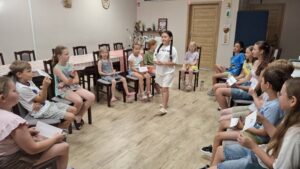 20260629_092651 Grupa dzieci siedzących w kole, oraz dziewczynka z długimi warkoczami stojąca po środku.