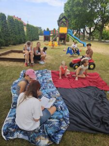 20260630_104156 Dziewczynka czyta książkę dzieciom siedzącym na placu zabaw.