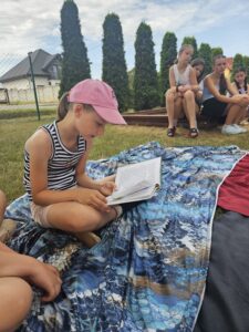 20260630_104231 Dziewczynka czyta książkę dzieciom siedzącym na placu zabaw.