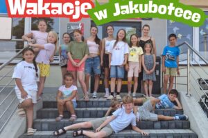 Grupa dzieci stojących na schodach przed biblioteką. Nad nimi napis: Wakacje w Jakubotece