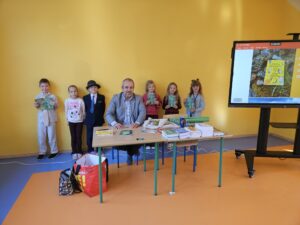 20260930_090809 Mężczyzna siedzący przy ławce, za nim stojące dzieci z książkami w rękach.